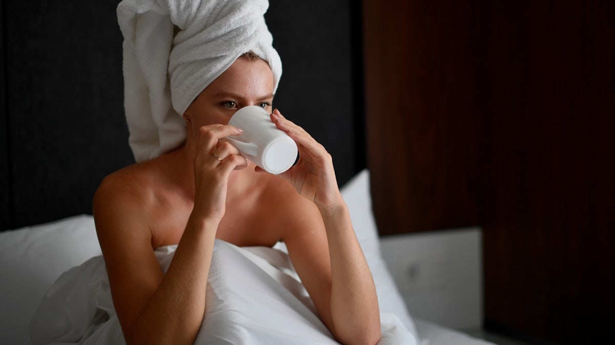 Young blonde woman with a white towel wrapped around her head drinking hot coffee in bed during her morning routine.